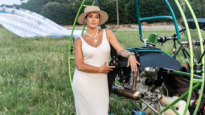 Femme en robe blanche posant près d'un paramoteur dans un champ.